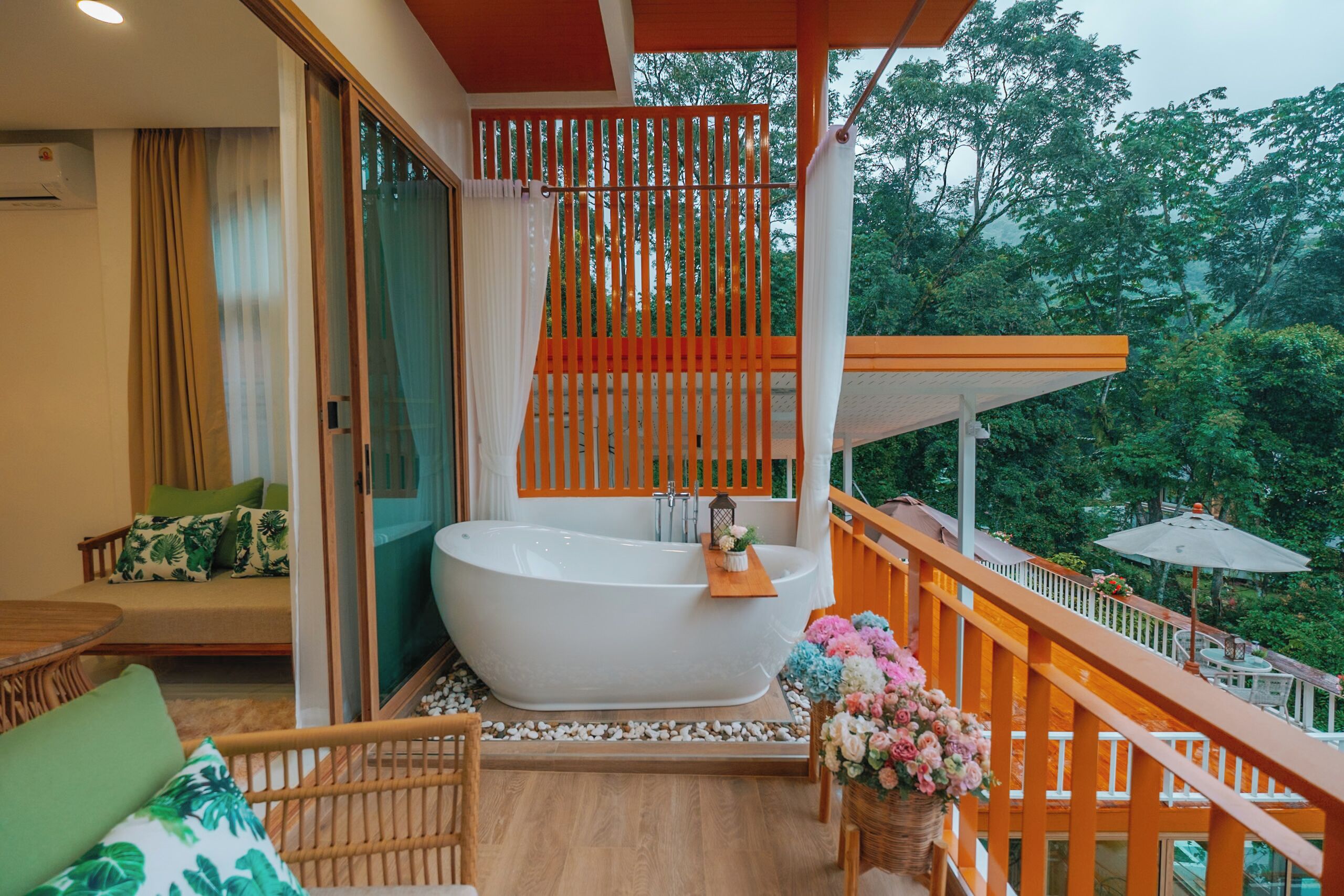 Un balcón con una bañera blanca, muebles de mimbre y vistas a árboles.