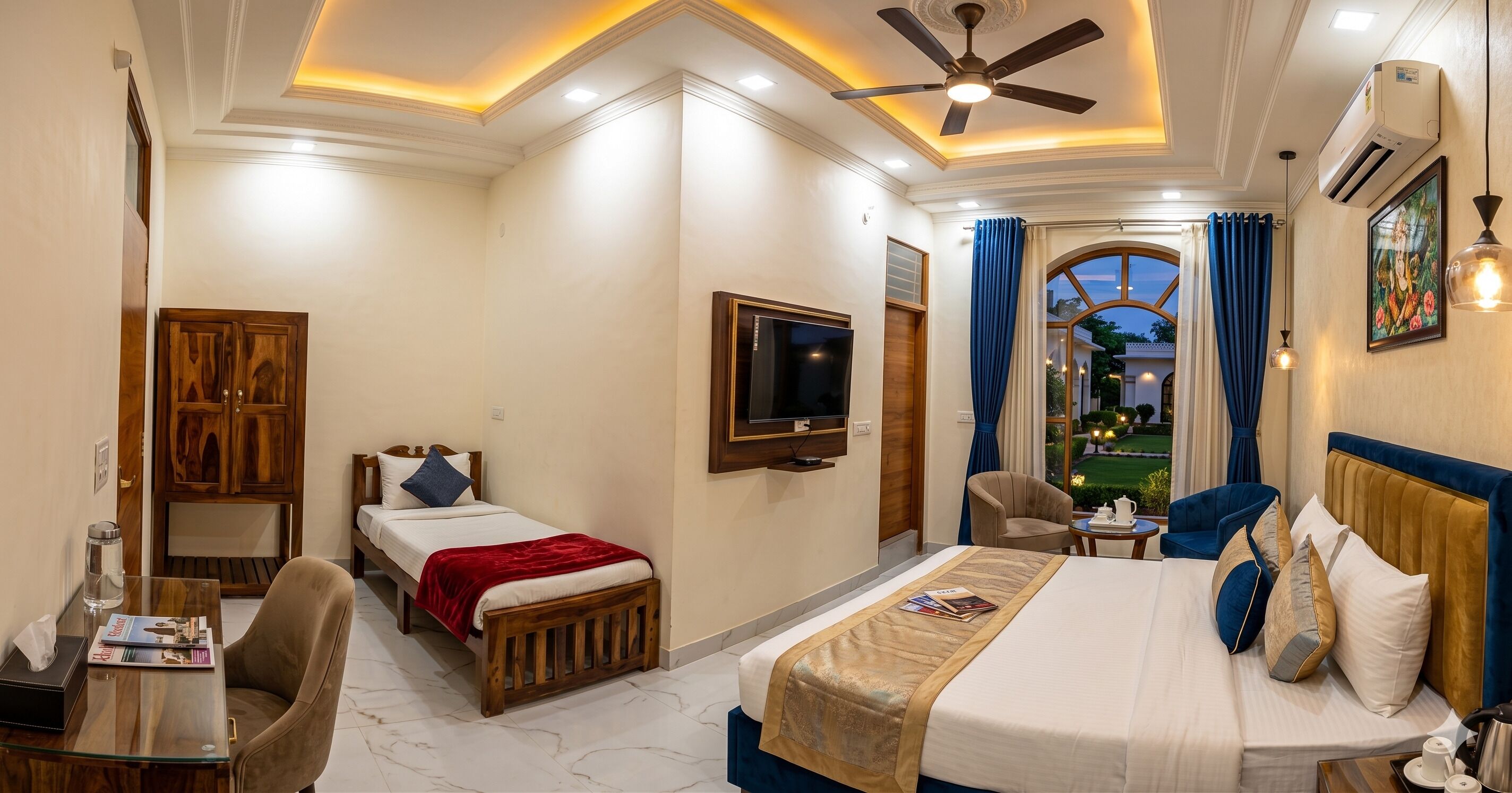 A hotel room with a bed, a TV, a desk with a chair, a window with curtains, and a ceiling fan.