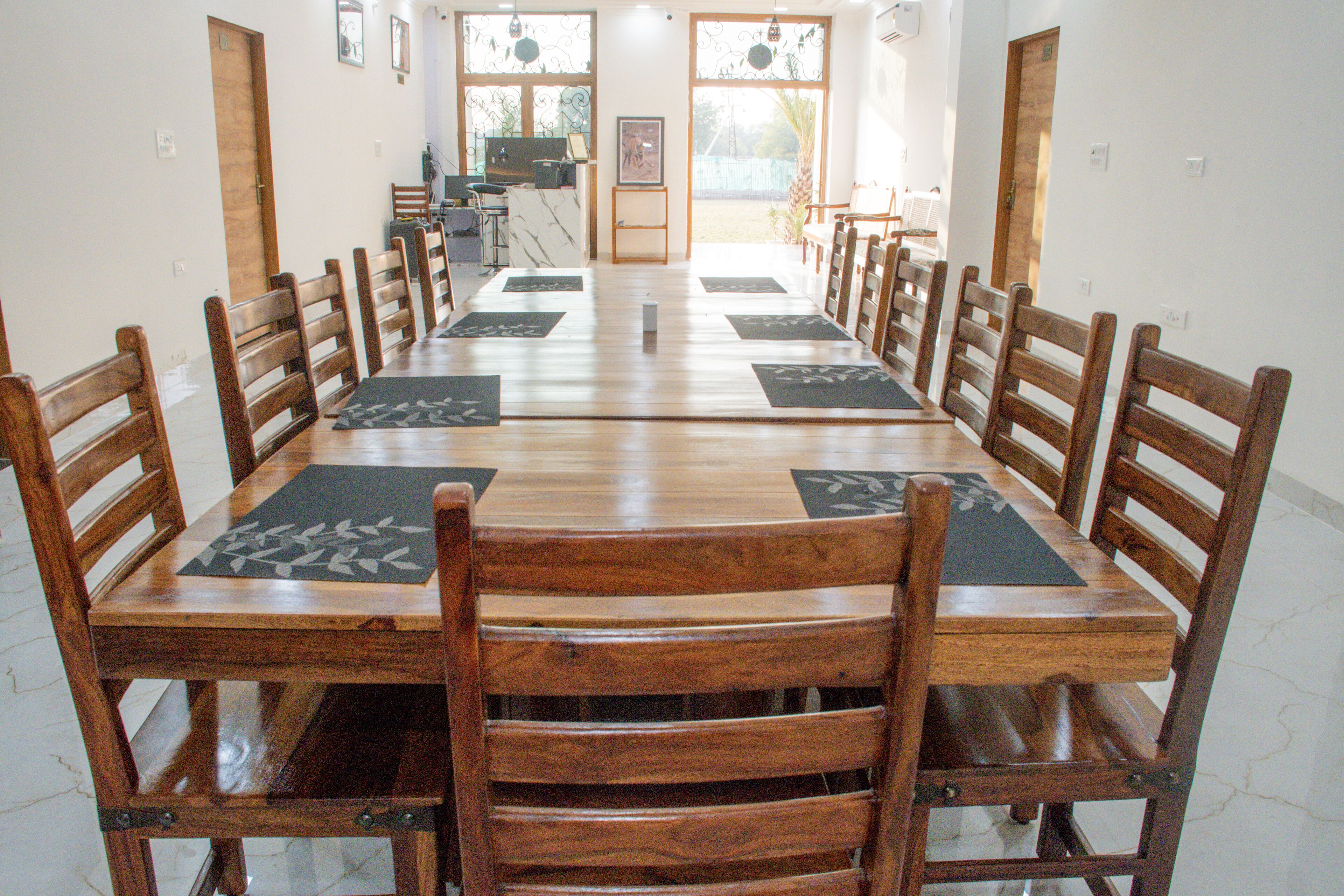 A long wooden dining table with matching chairs, a small kitchen area, and large windows with curtains.