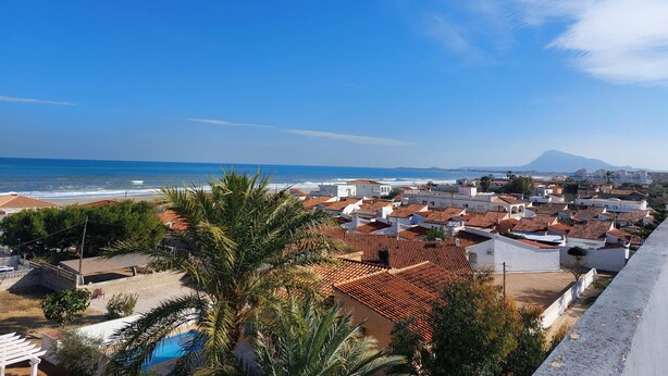 A coastal town with terracotta-roofed buildings, a beach, and a mountain in the distance.