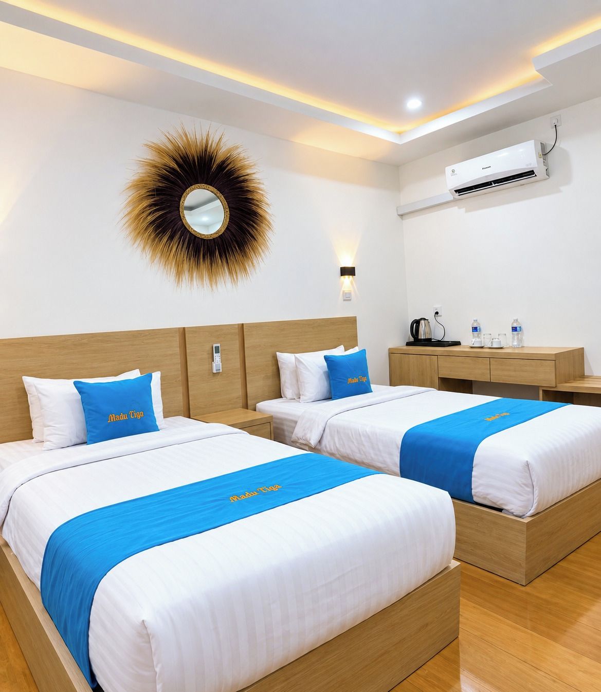 A hotel room with two beds, a wooden headboard, a wall-mounted air conditioner, a bedside table with a kettle, and a mirror with a sunburst design.