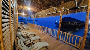 A wooden deck with lounge chairs overlooking a body of water, a thatched roof structure, and a wooden railing.
