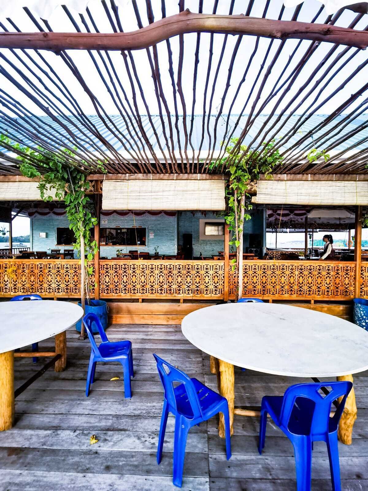 An outdoor seating area with wooden tables and blue plastic chairs, covered by a bamboo pergola.