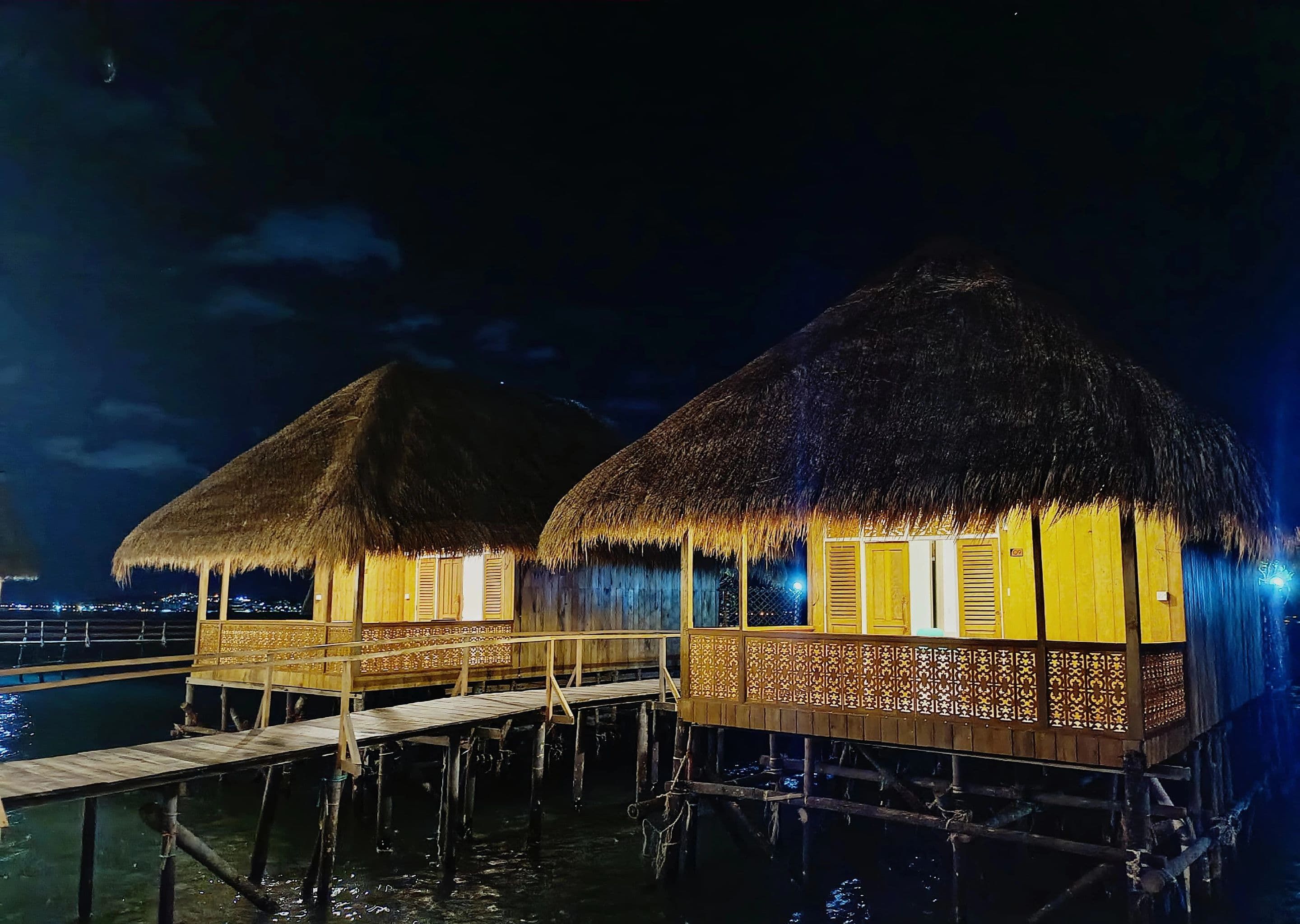 Bungalows sobre el agua con techos de paja y barandillas de madera, unidos por una pasarela de madera sobre el agua.