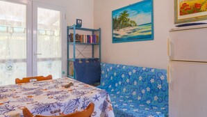 A room with a dining table, a blue sofa, a bookshelf, and a painting of a beach scene. - Apartment with garden in Lacona, ideal for relax. (Capoliveri)
