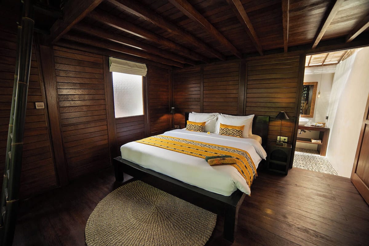 A wooden cabin bedroom with a bed, a nightstand, a window with a blind, and a bathroom visible through an open door.