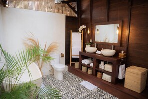 A bathroom with a white toilet, a white sink with a vessel basin, a wooden vanity with shelves, and a large mirror. - Kayu Village (Pererenan)