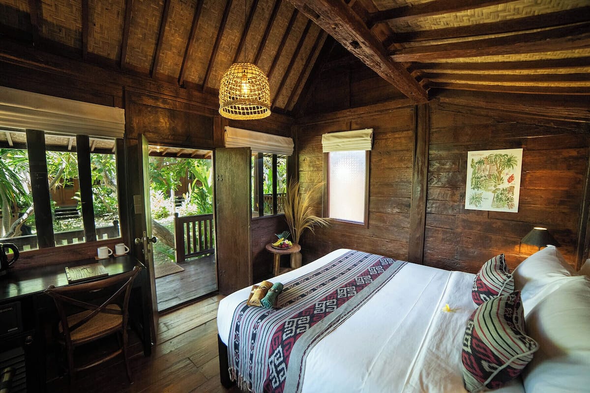 A rustic wooden room with a bed, a desk, and a window overlooking greenery.