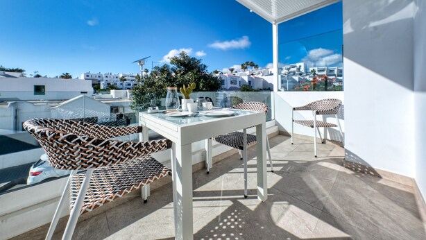 Eine Dachterrasse mit Tisch und Stühlen, einem Kaktus und Blick auf weiße Gebäude unter blauem Himmel.