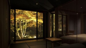 Una habitación con suelo de tatami, una mesa baja, un sofá y un ventanal con vistas a árboles.