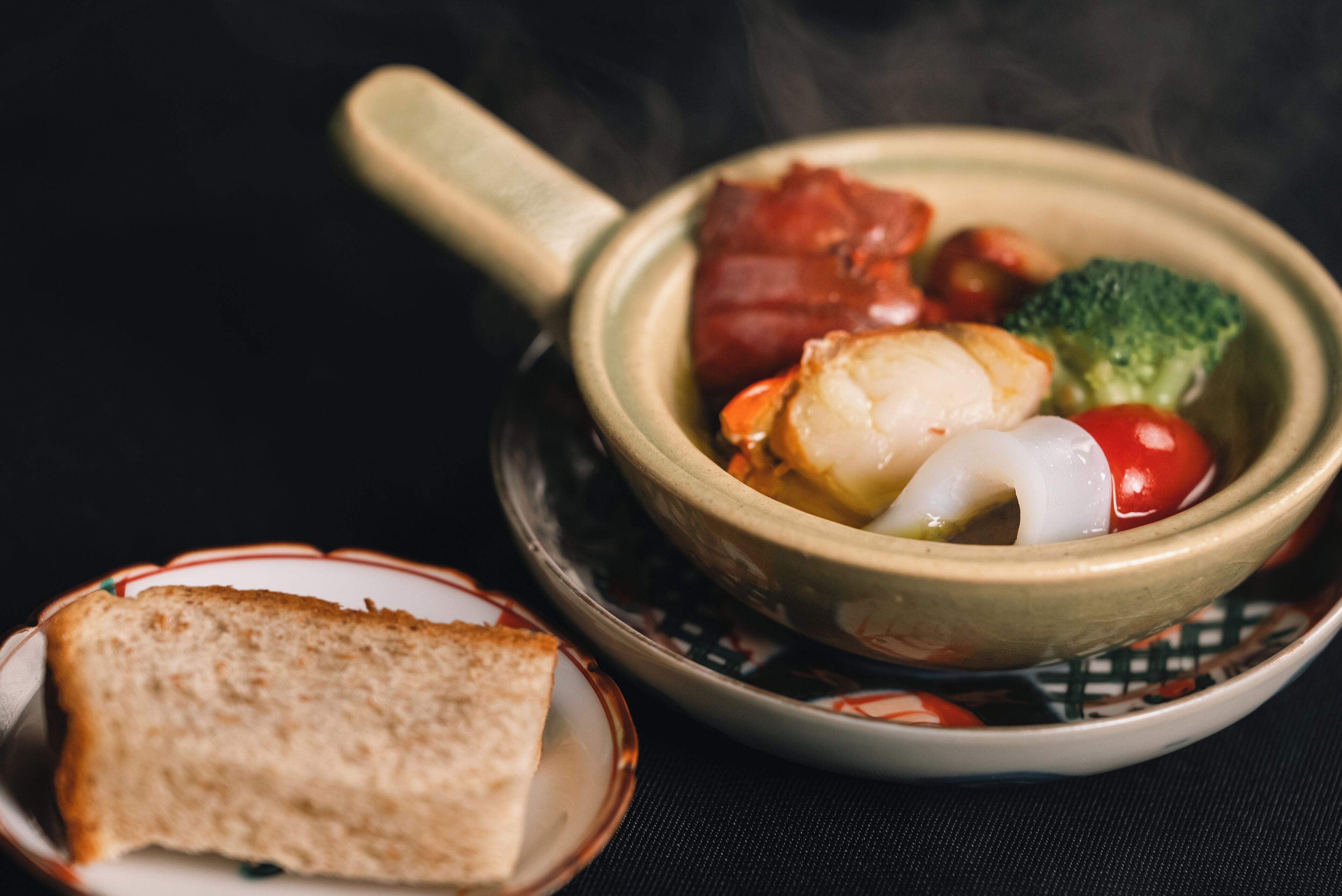 A bowl of food with a handle, containing various vegetables and meat, accompanied by a slice of bread on a separate plate.
