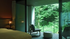 Una habitación de hotel moderna con una cama grande, un área de descanso con una silla y reposapiés, y un balcón con vistas a vegetación exuberante.