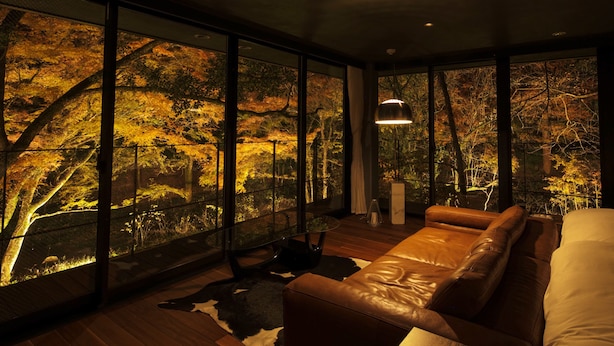 Una habitación moderna con un amplio ventanal que da a un bosque. Hay un sofá de cuero y una cama con cabecera blanca.