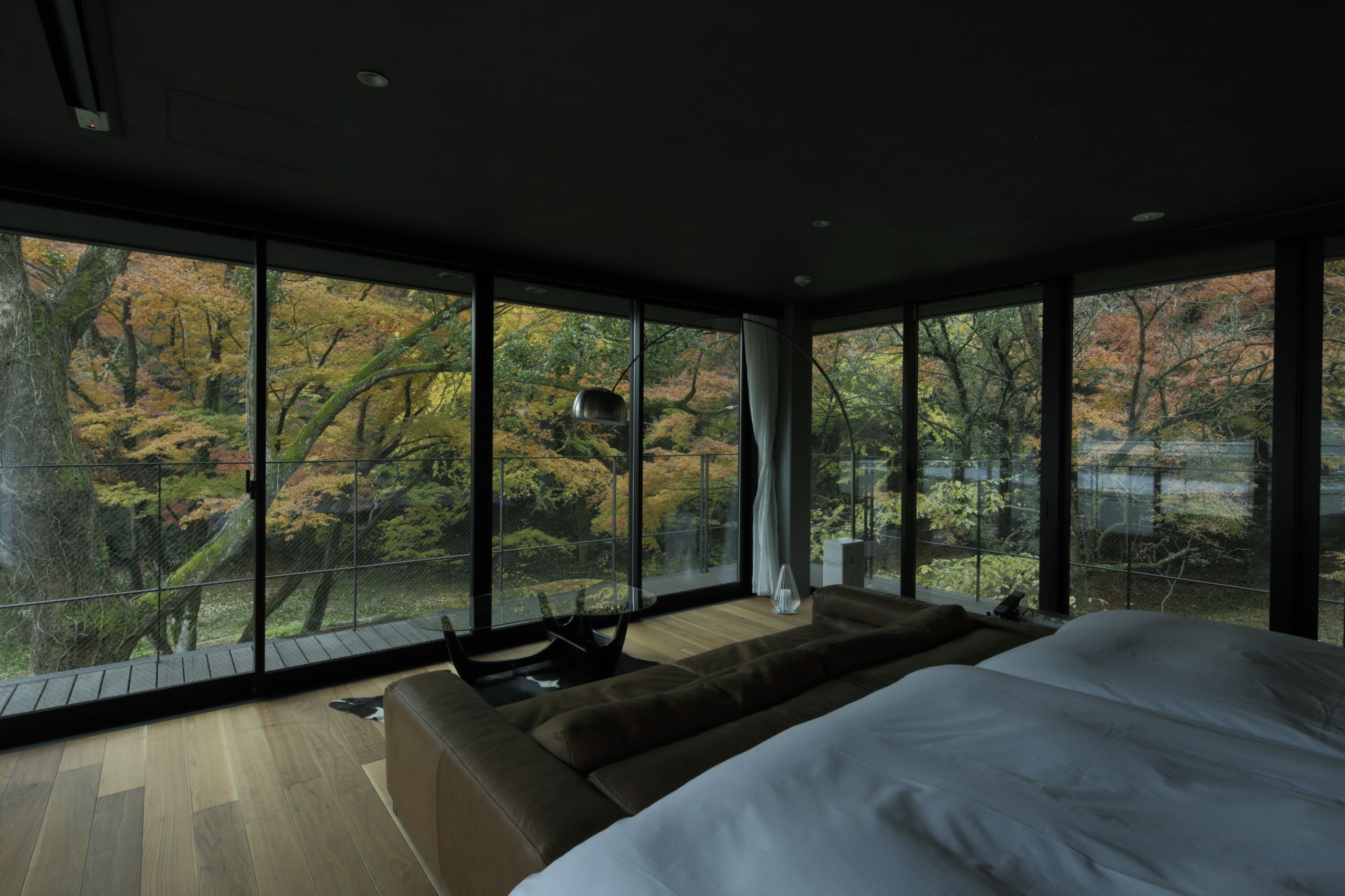 A modern bedroom with a large bed, a sofa, and a view of autumn trees through floor-to-ceiling windows.