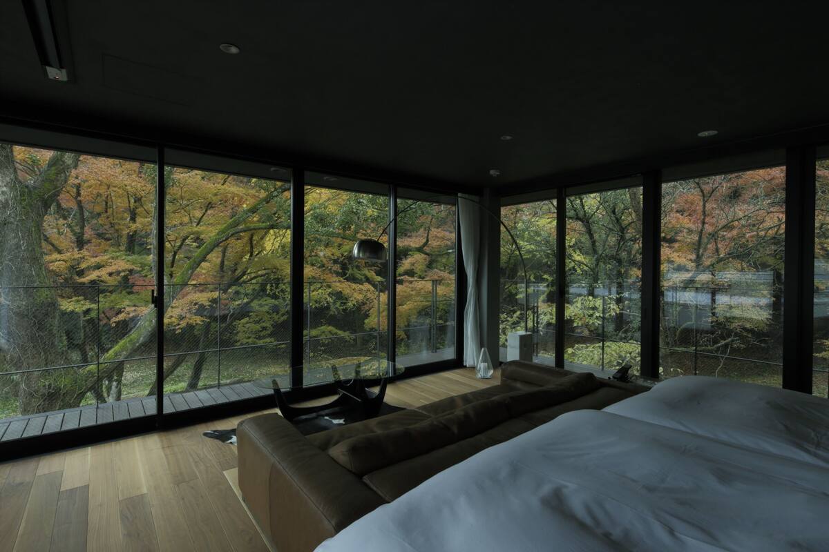 Une chambre moderne avec un grand lit, un canapé et une vue sur les arbres d’automne grâce à de grandes fenêtres du sol au plafond.