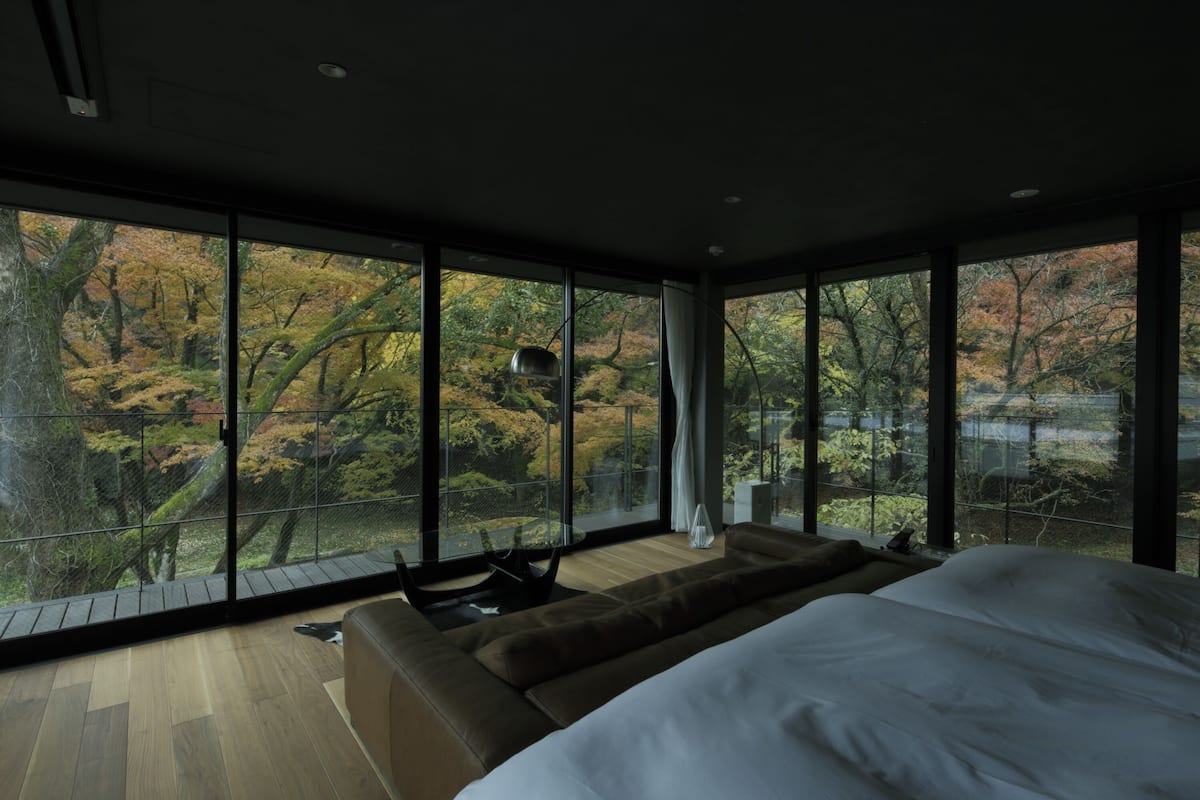 A modern bedroom with a large bed, a sofa, and a view of autumn trees through floor-to-ceiling windows.