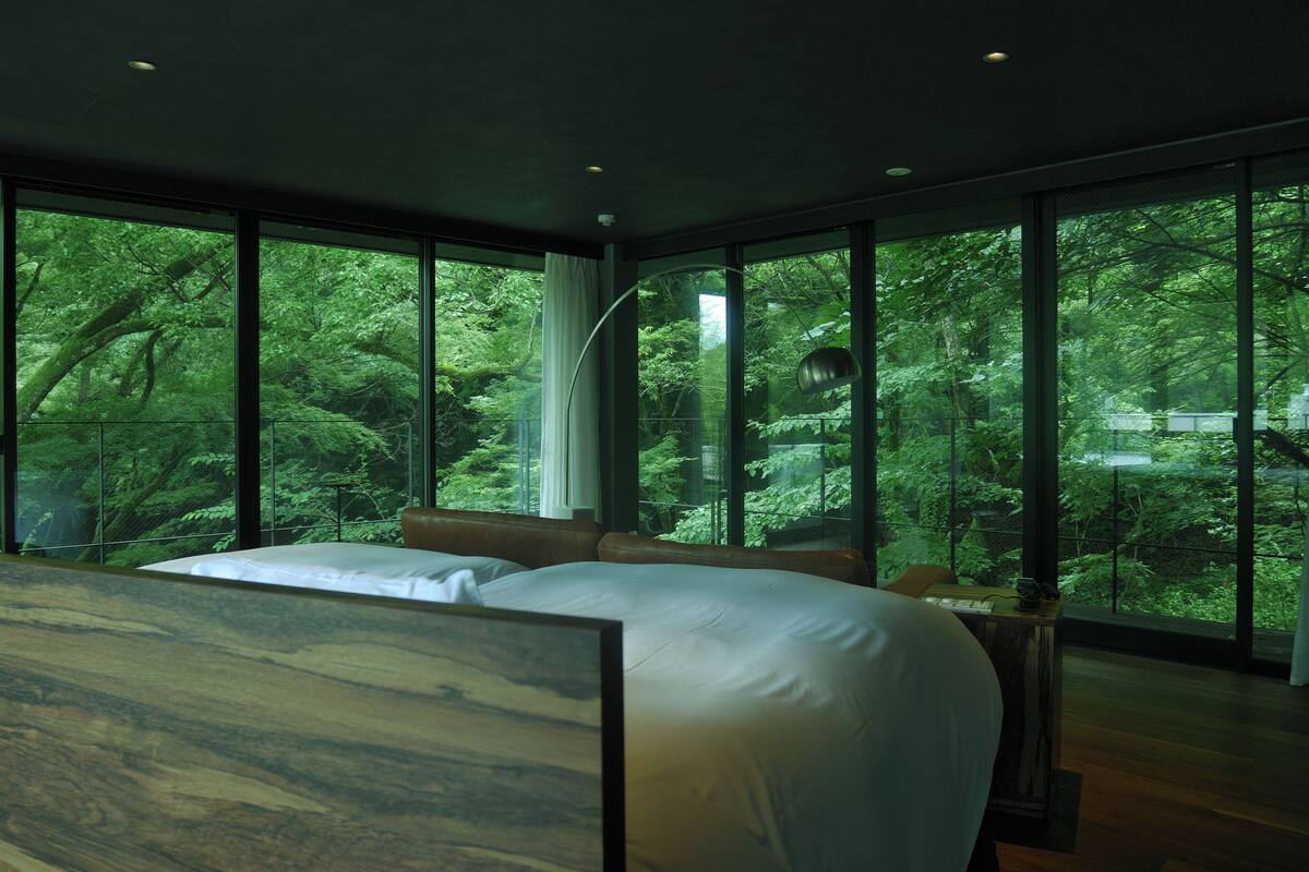 Une chambre avec un grand lit, un bureau et une vue sur une forêt, grâce à de grandes fenêtres.