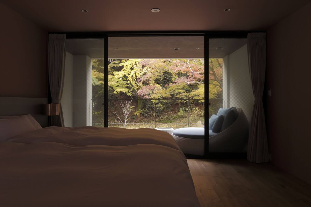 Une chambre avec un grand lit, une chaise et une vue sur les arbres, visible à travers une porte coulissante.