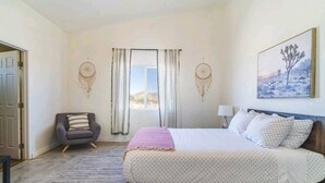 A bedroom with a bed, a chair, a window with curtains, and a wall-mounted dreamcatcher.
