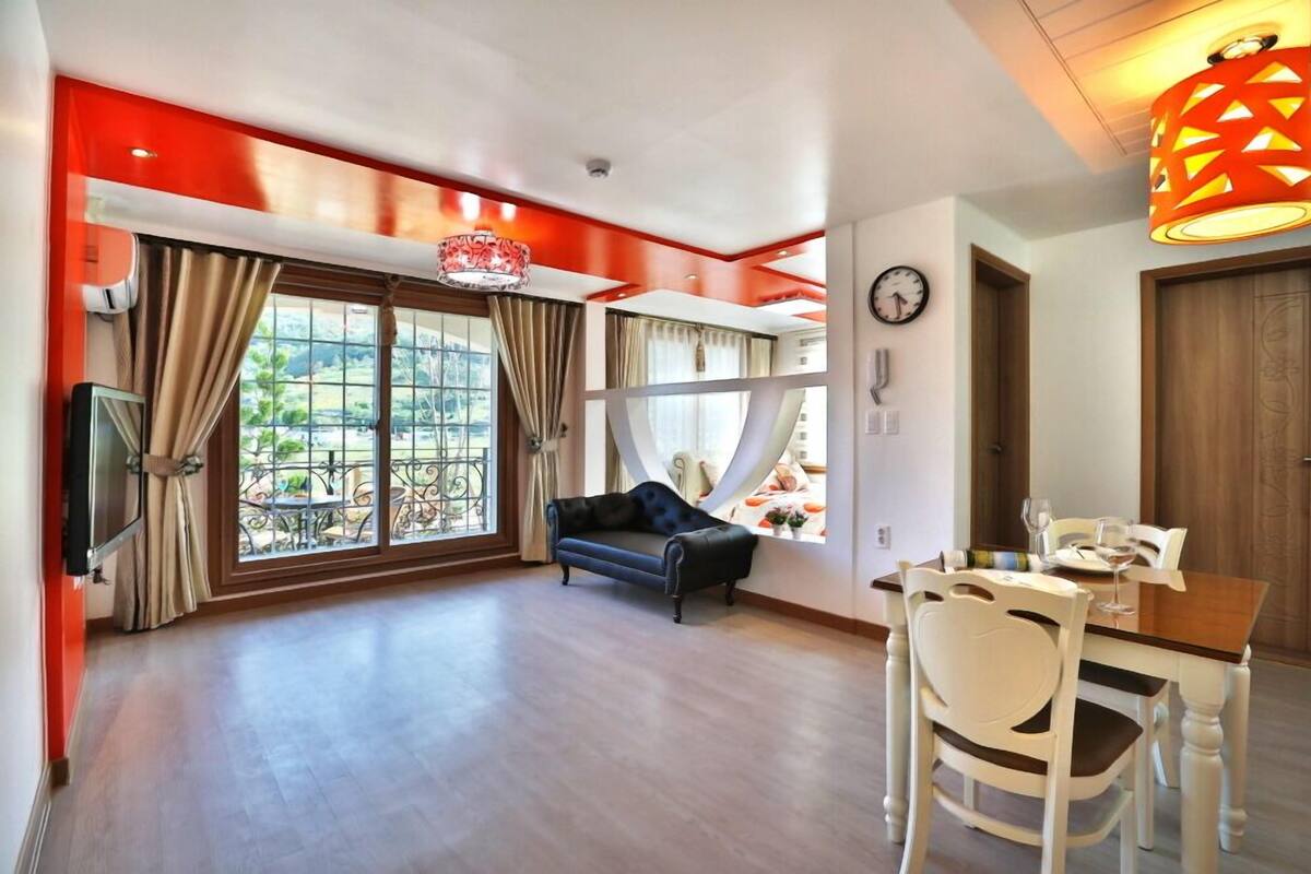 A spacious living room with a large window, a sofa, a dining table with chairs, and a decorative hanging light fixture.