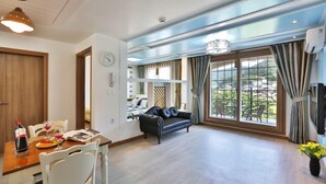 A spacious living area with a large window, a black leather sofa, and a dining table set with a bottle of wine and glasses. - Monami Pension (Tongyeong)