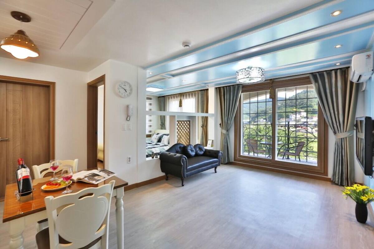 A spacious living area with a large window, a black leather sofa, and a dining table set with a bottle of wine and glasses.