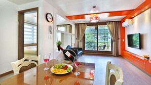 A dining area with a table set for two, a bottle of wine, and a bowl of fruit. There is a TV mounted on the wall and a window with curtains. - Monami Pension (Tongyeong)