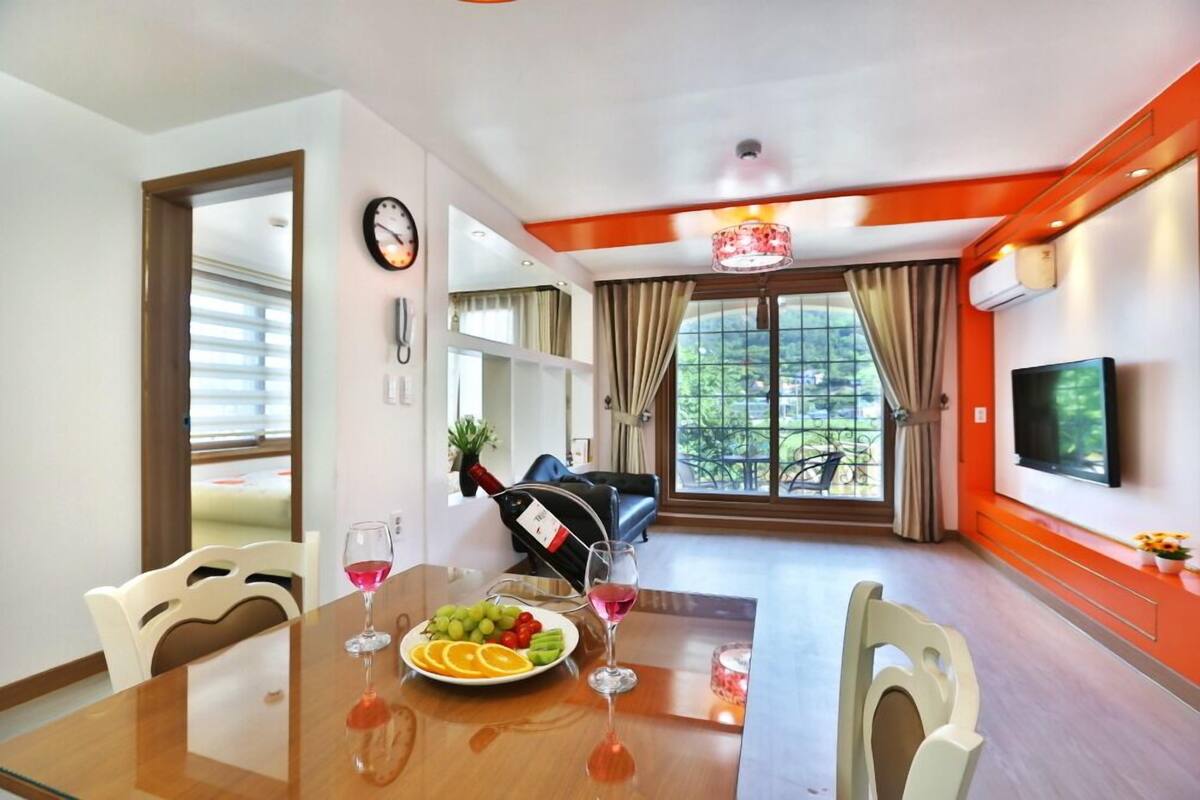 A dining area with a table set for two, a bottle of wine, and a bowl of fruit. There is a TV mounted on the wall and a window with curtains.