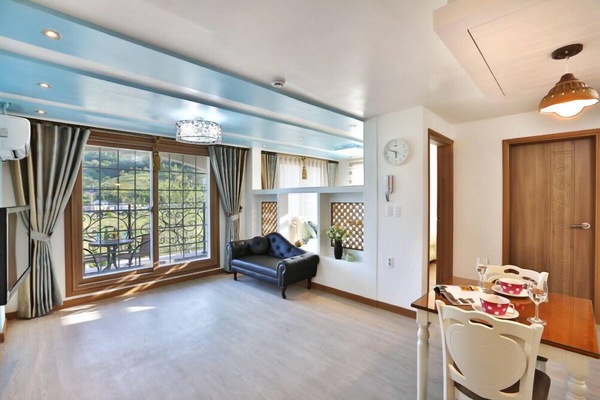 A spacious room with a large window, a sofa, a dining table with chairs, and a wooden door.