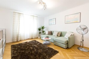 A modern living room with a green sofa, a round coffee table, a large rug, and a standing fan.