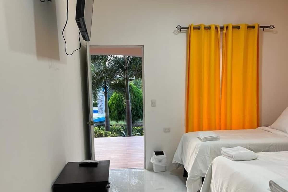 A hotel room with two beds, a TV mounted on the wall, a small black cabinet, and a window with a view of greenery outside.