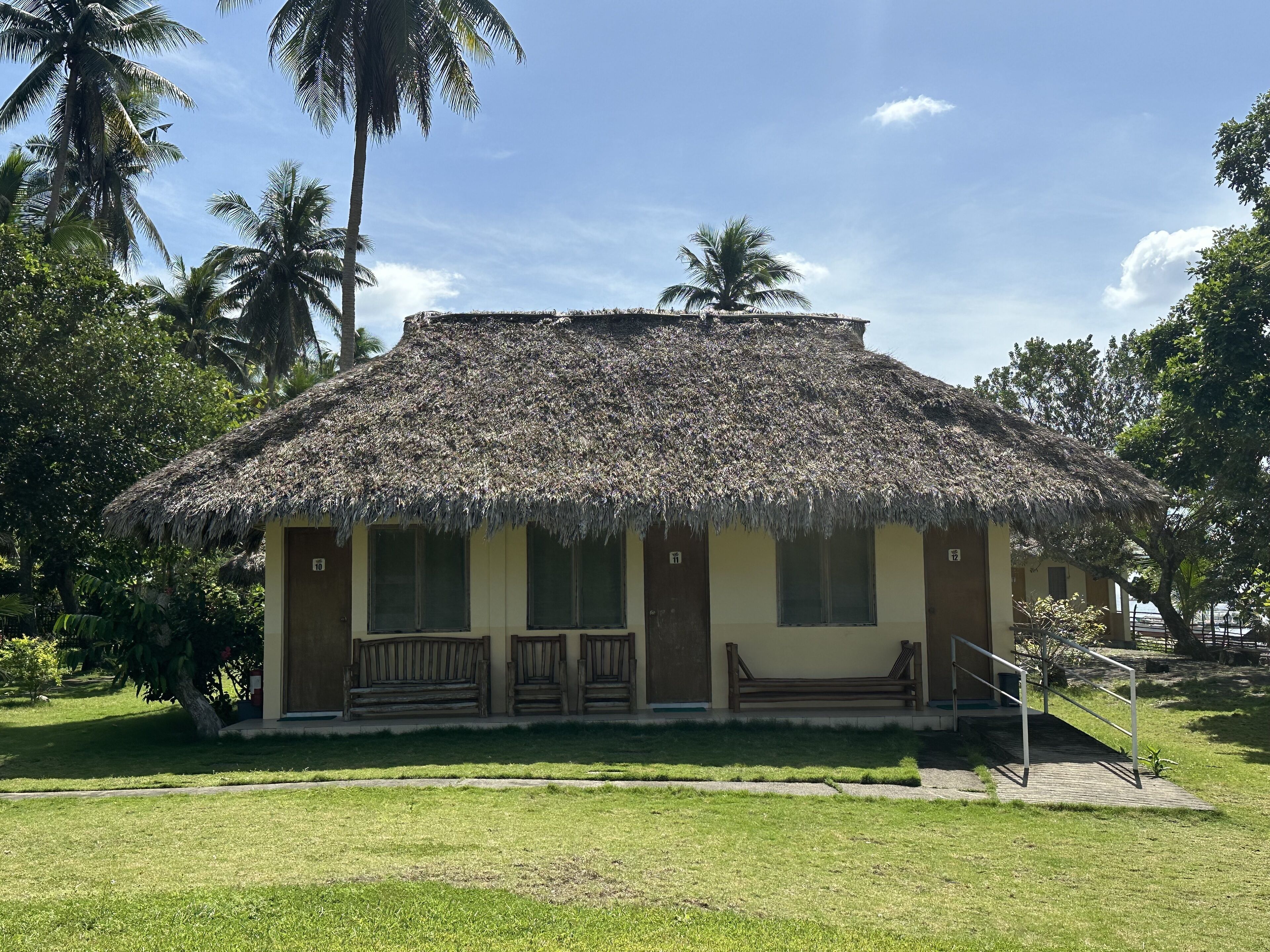 A thatched-roof building with a porch and benches, surrounded by lush greenery and palm trees.