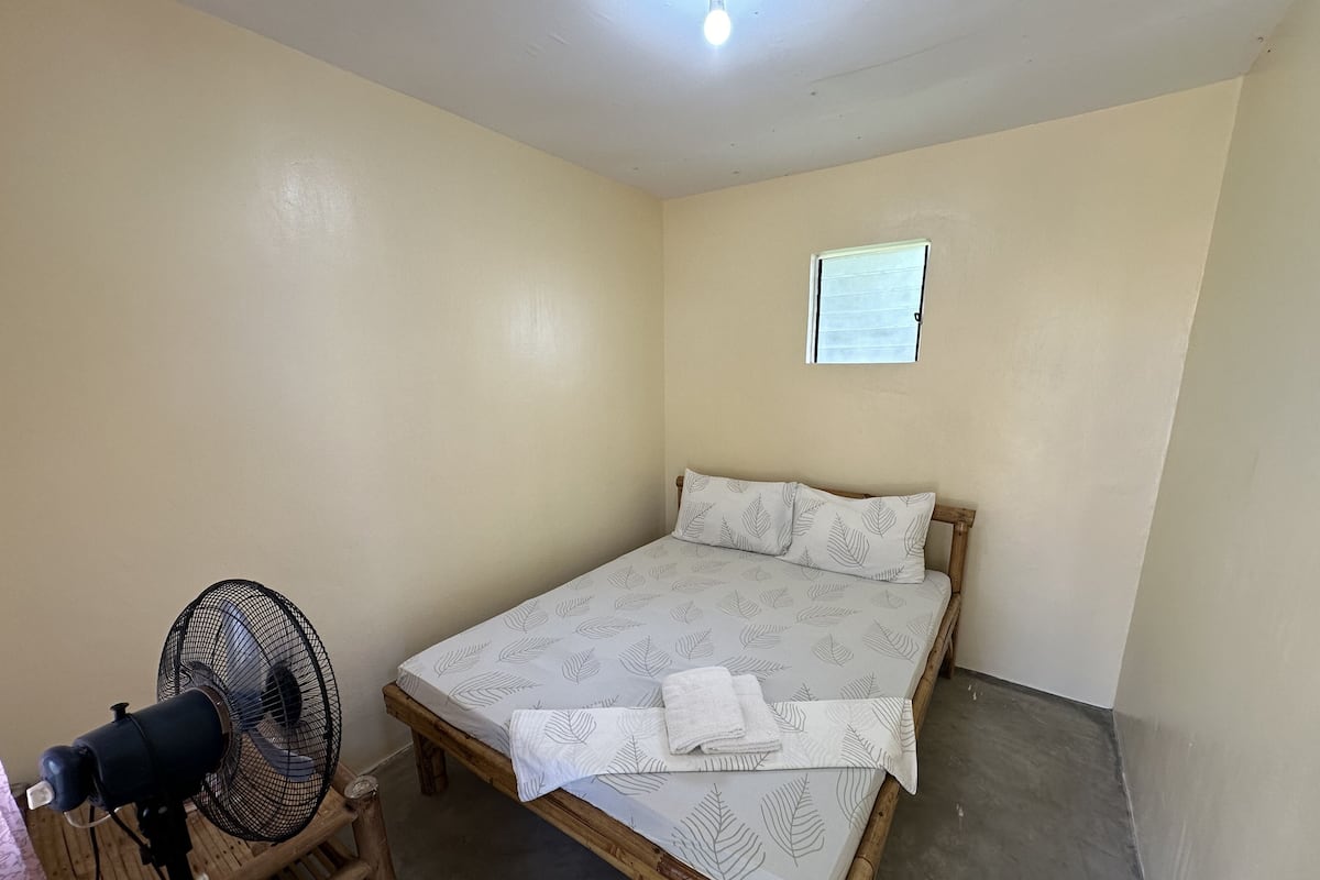 A small, single-bed room with a wooden frame, a white mattress with leaf patterns, two pillows, a standing fan, and a small window.