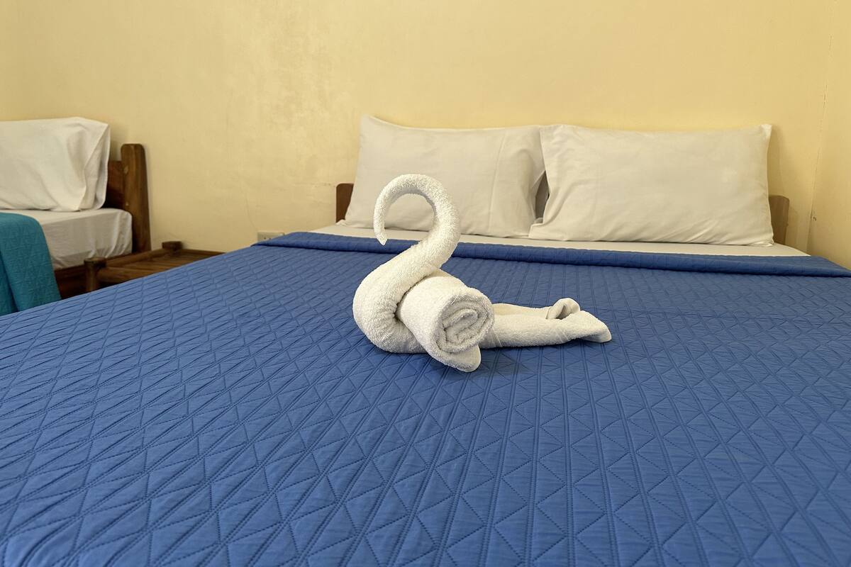 A bed with a blue quilted bedspread and a white swan-shaped towel on it.