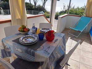 Uma mesa posta com pratos, um jarro e uma tigela de frutas, com vista para o mar ao fundo.