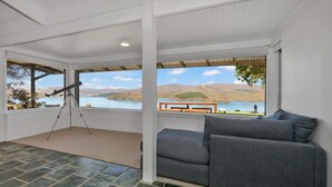 Habitació amb una finestra gran que ofereix una vista panoràmica d'un llac i muntanyes. Hi ha un telescopi col·locat al costat esquerre, i un sofà gris amb coixins a la dreta.