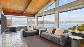 A spacious room with a large window offering a scenic view of a lake and hills. There are two grey sofas with cushions and a small round table. - Lake Eildon Retreat - Breathtaking Views - Sleeps 12 (Mansfield)