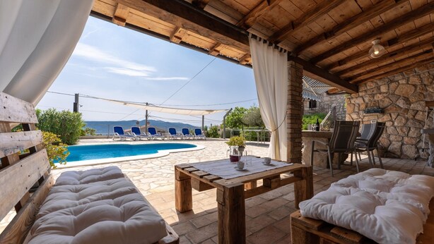 A wooden outdoor seating area with cushioned benches, a stone table, and a view of a swimming pool and mountains. - Villa Zaria (Kvarner - Mošćenička Draga) (Mošćenička Draga)