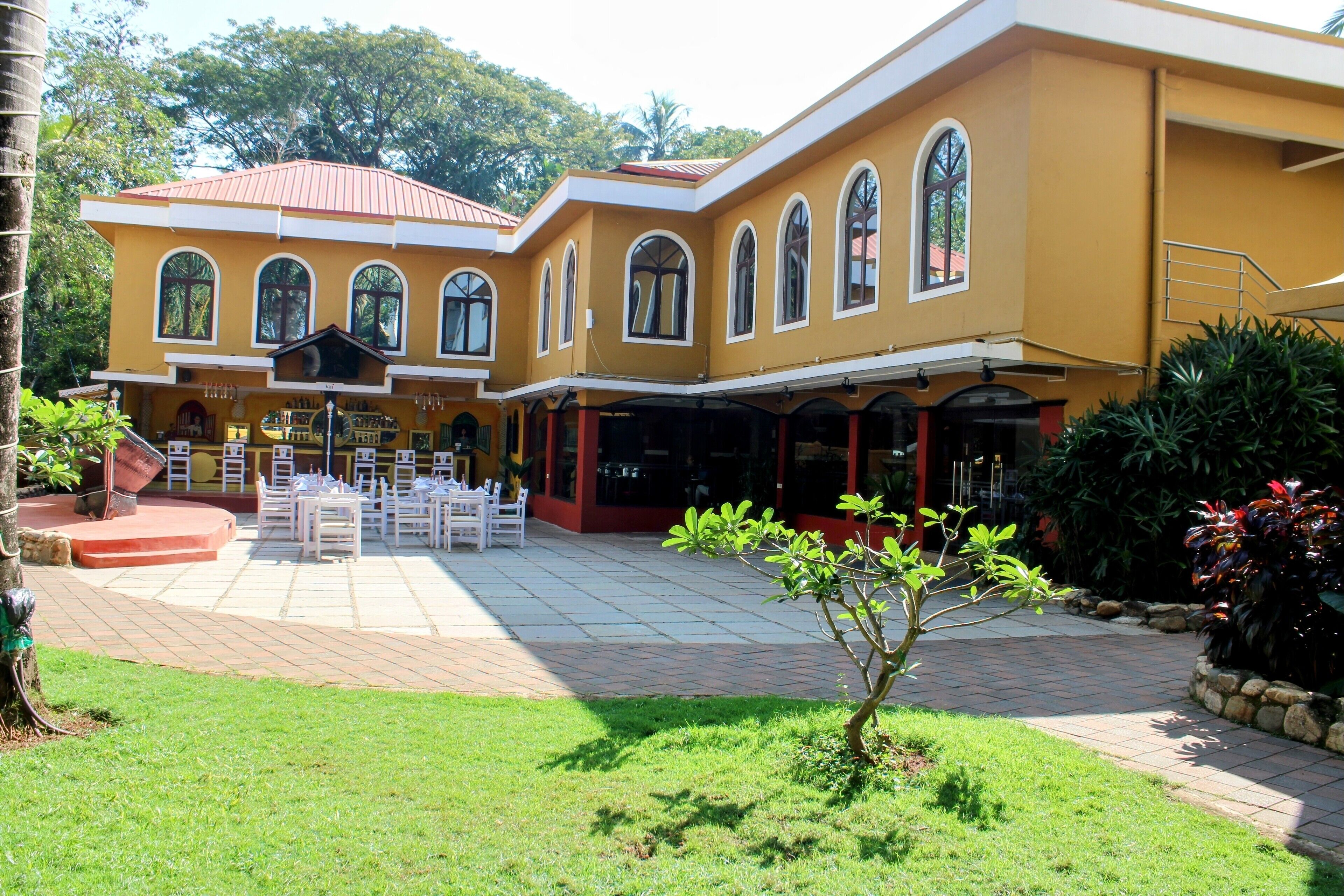 A yellow building with arched windows and a red roof, featuring a patio area with white tables and chairs.