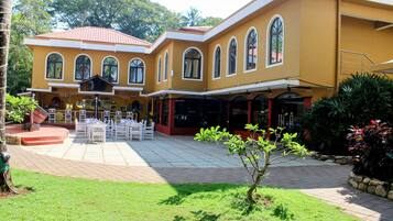 A yellow building with arched windows and a red roof, featuring a patio area with white tables and chairs.