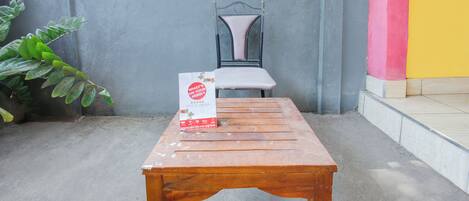 Una mesa de madera con un letrero encima, una silla de metal, una planta en maceta y una pared con una vista parcial de un edificio colorido.