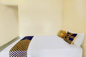 A hotel room with a single bed, a patterned bedspread, two pillows with geometric designs, and a plain wall.