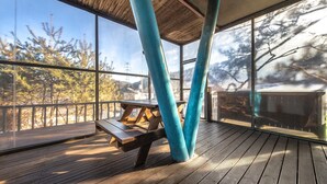 Una terraza de madera con una mesa de picnic, ventanas amplias y vistas a árboles y montañas.