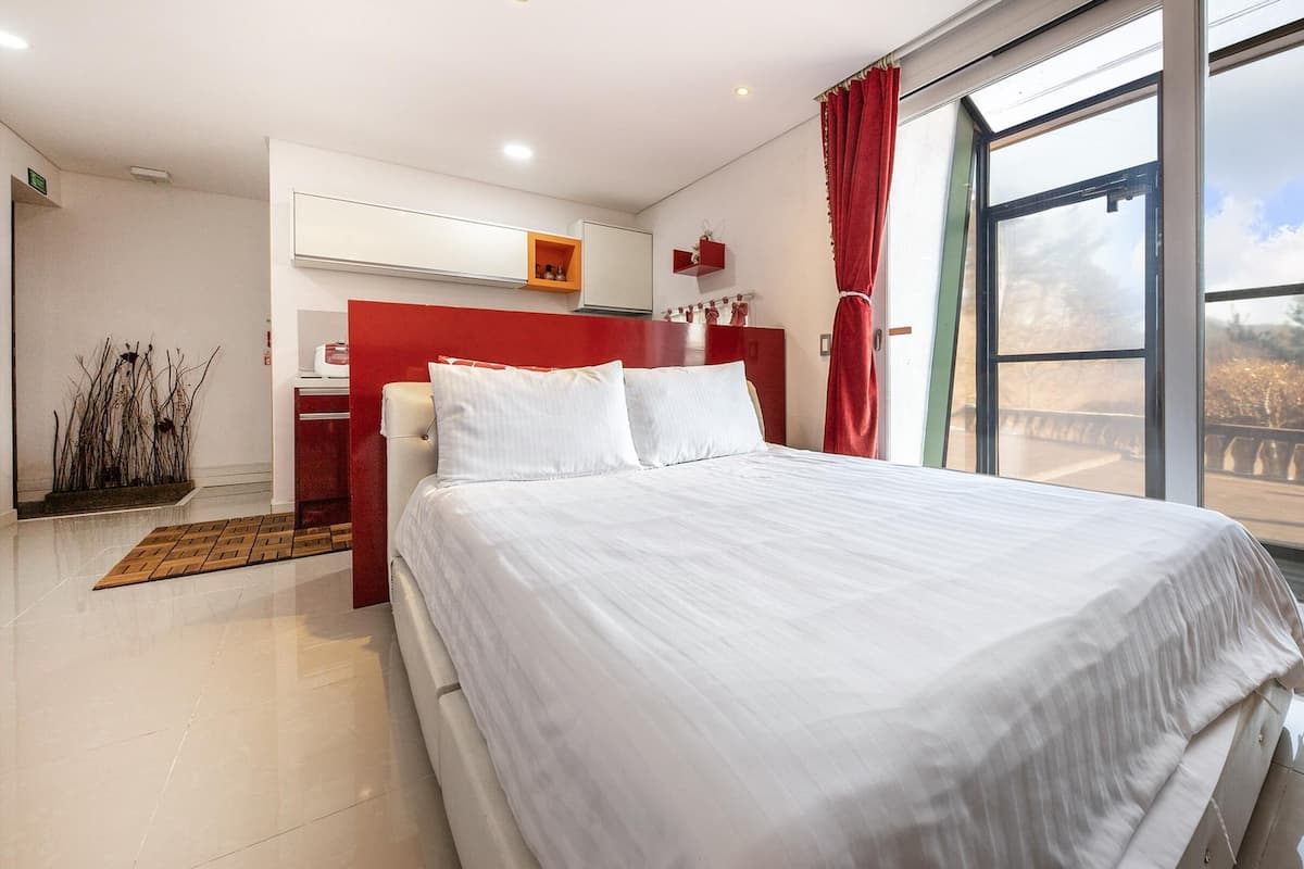 Une chambre d’hôtel moderne avec un grand lit, un mur d’accent rouge et une vue sur un pont et des arbres, visible par la fenêtre.