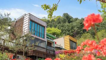 Edifici modern amb grans finestres i un balcó, envoltat de vegetació exuberant i plantes florejants.
