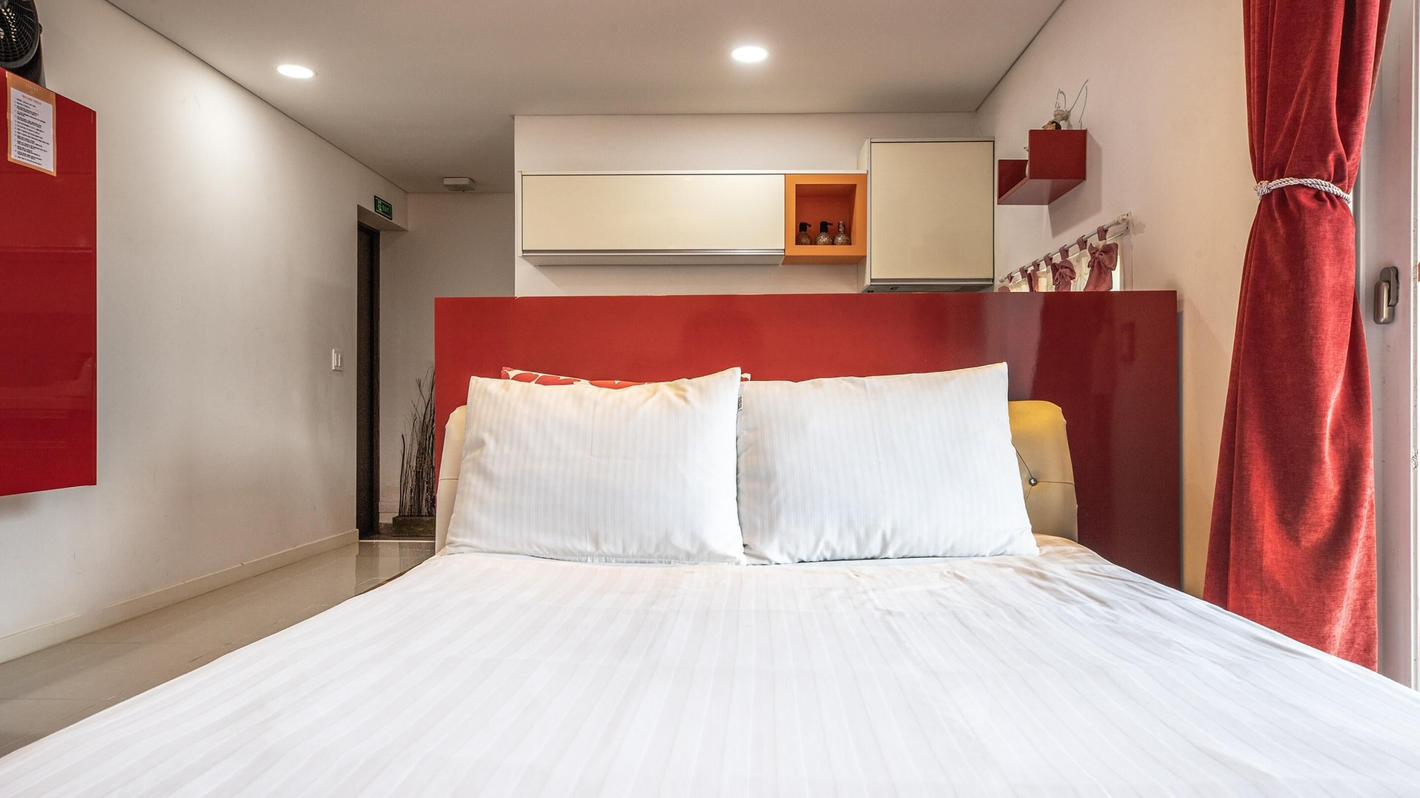 A hotel room with a bed, a red accent wall, a wardrobe, and a small shelf with decorative items.