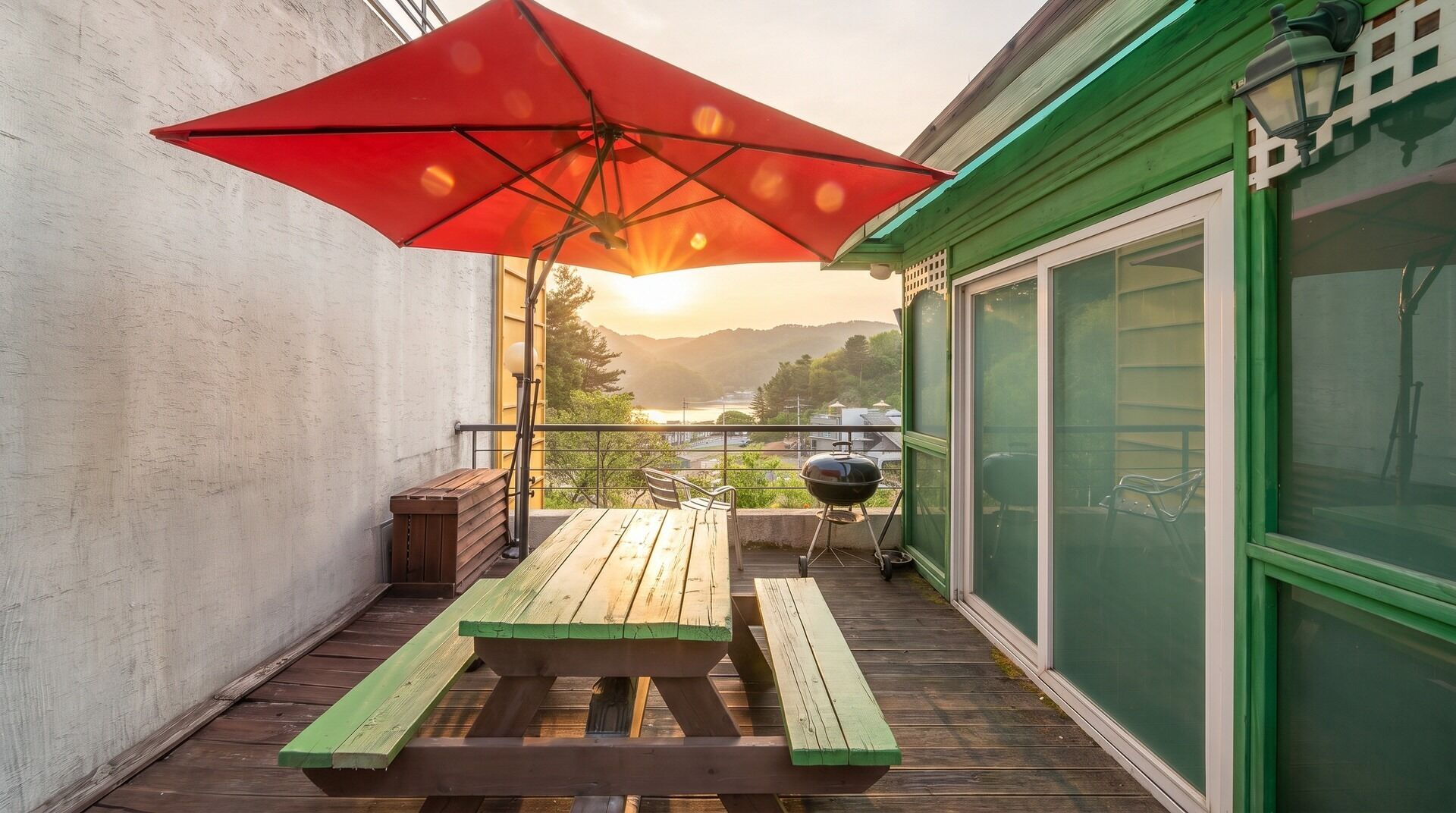 A wooden deck with a picnic table and a red umbrella, overlooking a scenic view with mountains and a body of water.