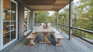 An outdoor patio with a wooden table and benches, surrounded by glass windows and a view of trees. - Villa in Durbuy near Golf Course (Durbuy)