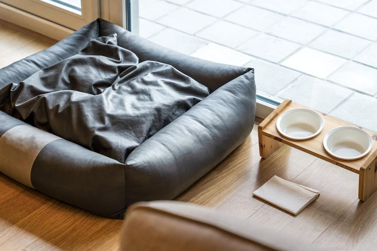 A pet bed with a cushion and two bowls on a wooden stand.