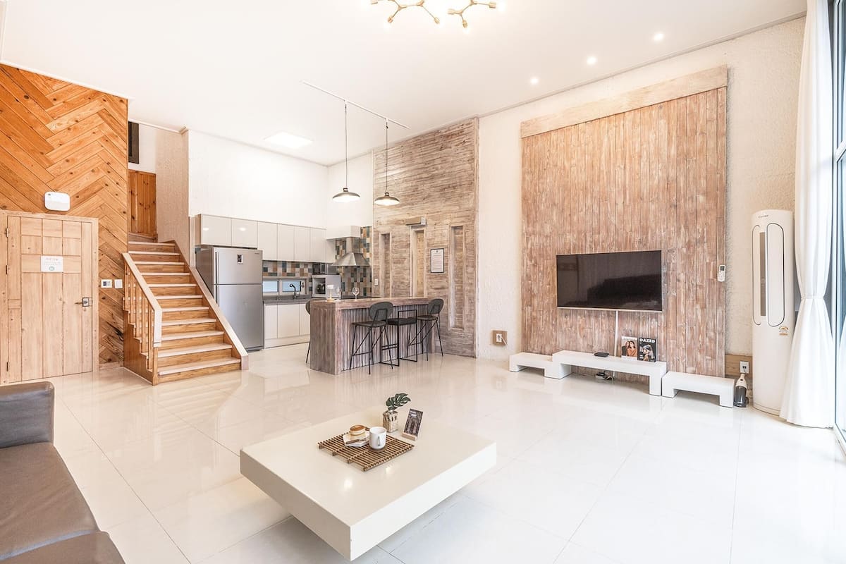 A modern living room with a white coffee table, a kitchen with white cabinets, a wooden staircase, and a large TV mounted on a wooden wall.